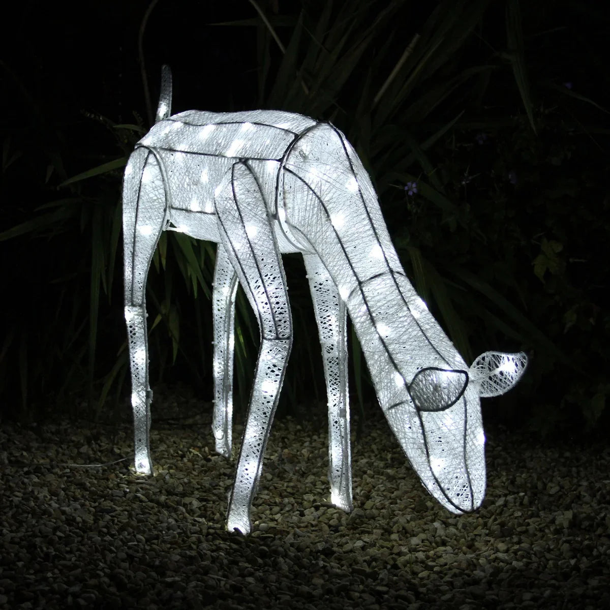 Glowing white light-up doe sculpture with intricate mesh design, set against a dark background of greenery and pebbles
