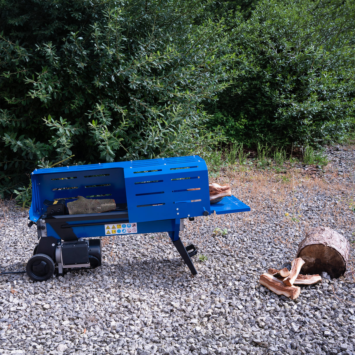 Bright blue electric log splitter on gravel, with split logs nearby and greenery in the background, showcasing its sturdy design and functionality