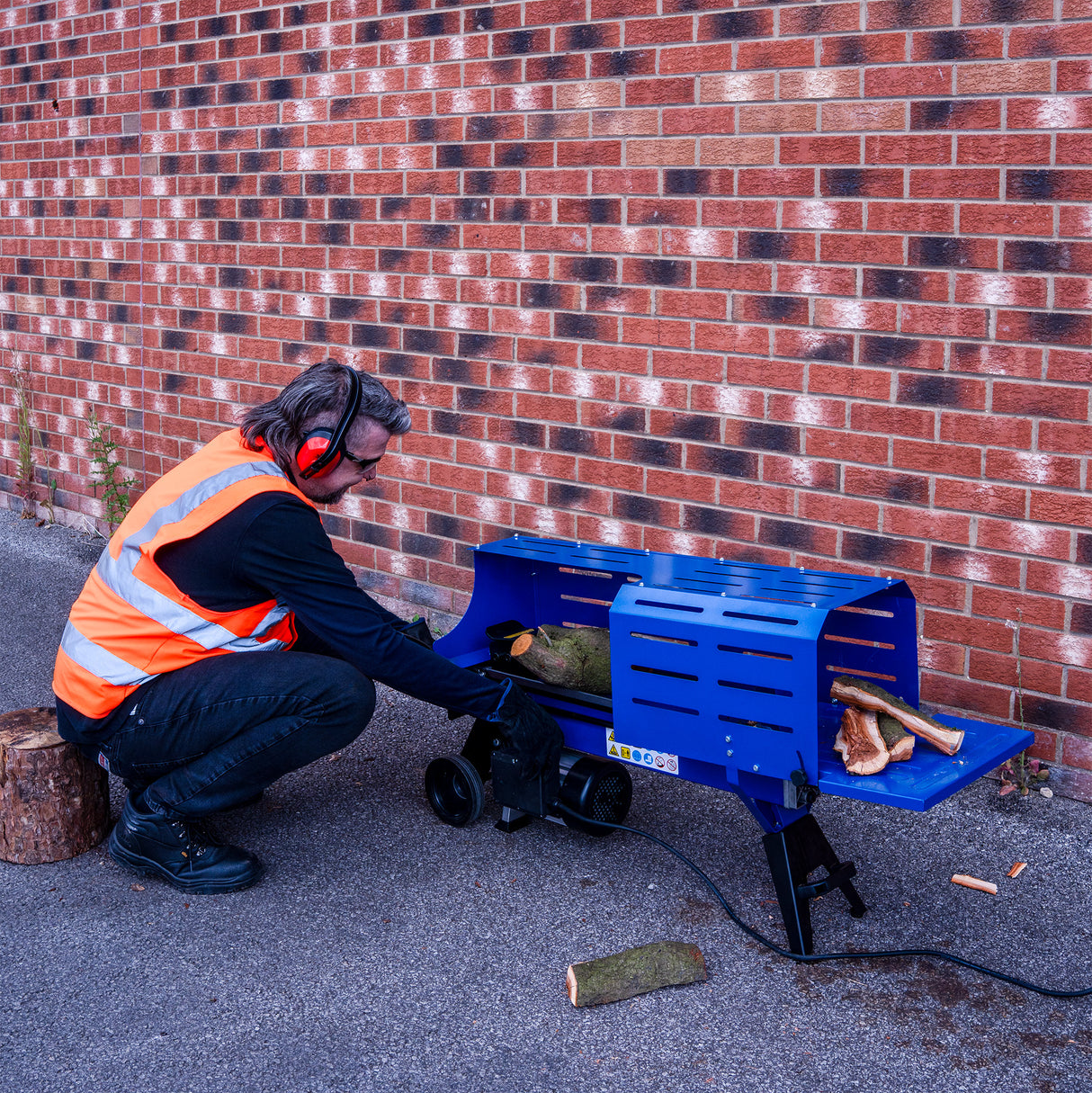 Electric log splitter in blue, with a person in an orange safety vest operating it, splitting a log against a brick wall background