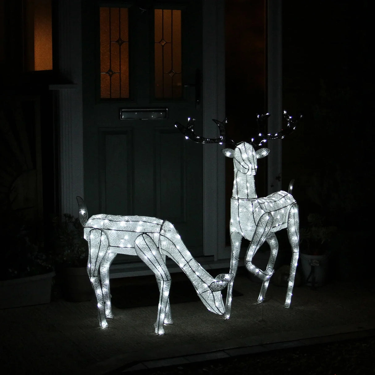 Large light-up stag and doe reindeer set with white wireframe, glowing LED lights, standing near a front door in evening light