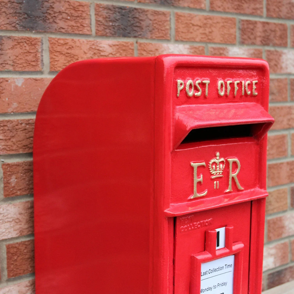 Bright red Royal Mail post box with gold lettering, featuring a curved top and a collection slot, set against a brick wall