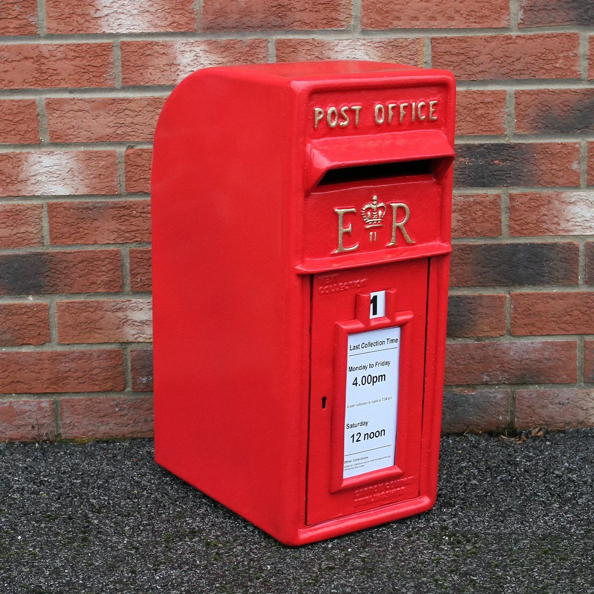 Bright red Royal Mail post box with a textured finish, featuring a crown emblem and collection time notice against a brick wall