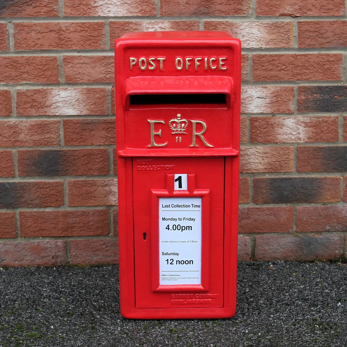 Bright red Royal Mail post box with stand, featuring gold lettering, a collection slot, and a notice for collection times against a brick wall