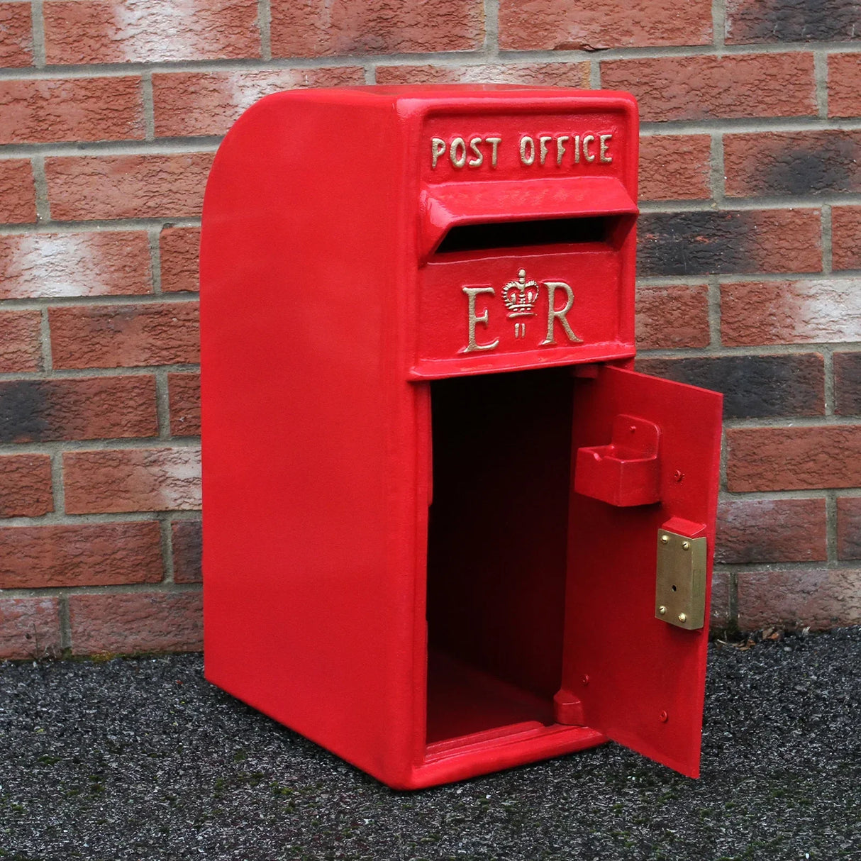 Bright red Royal Mail post box with gold lettering, featuring an open door, set against a brick wall background