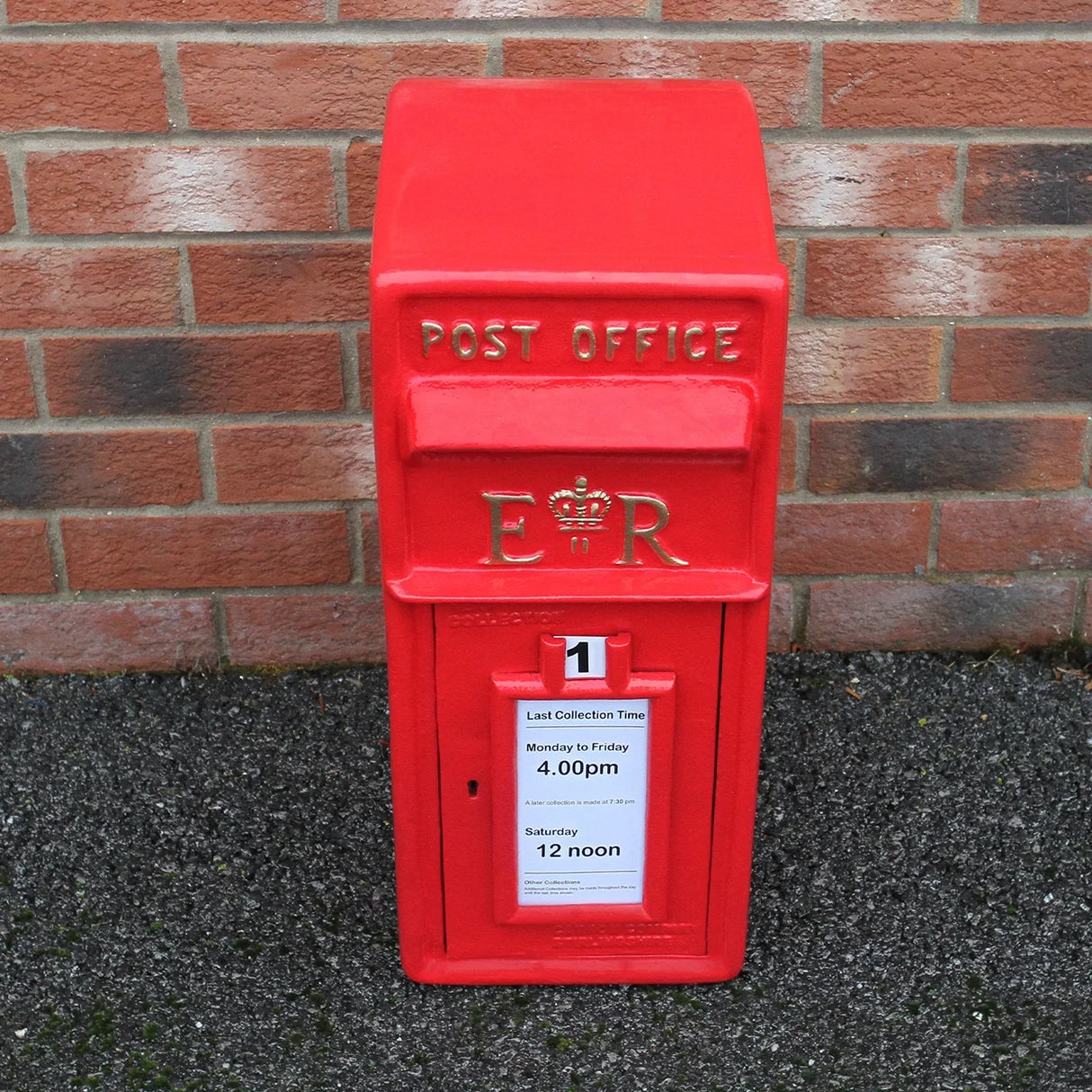 Bright red Royal Mail post box with gold lettering, featuring collection times and a classic design, set against a brick wall