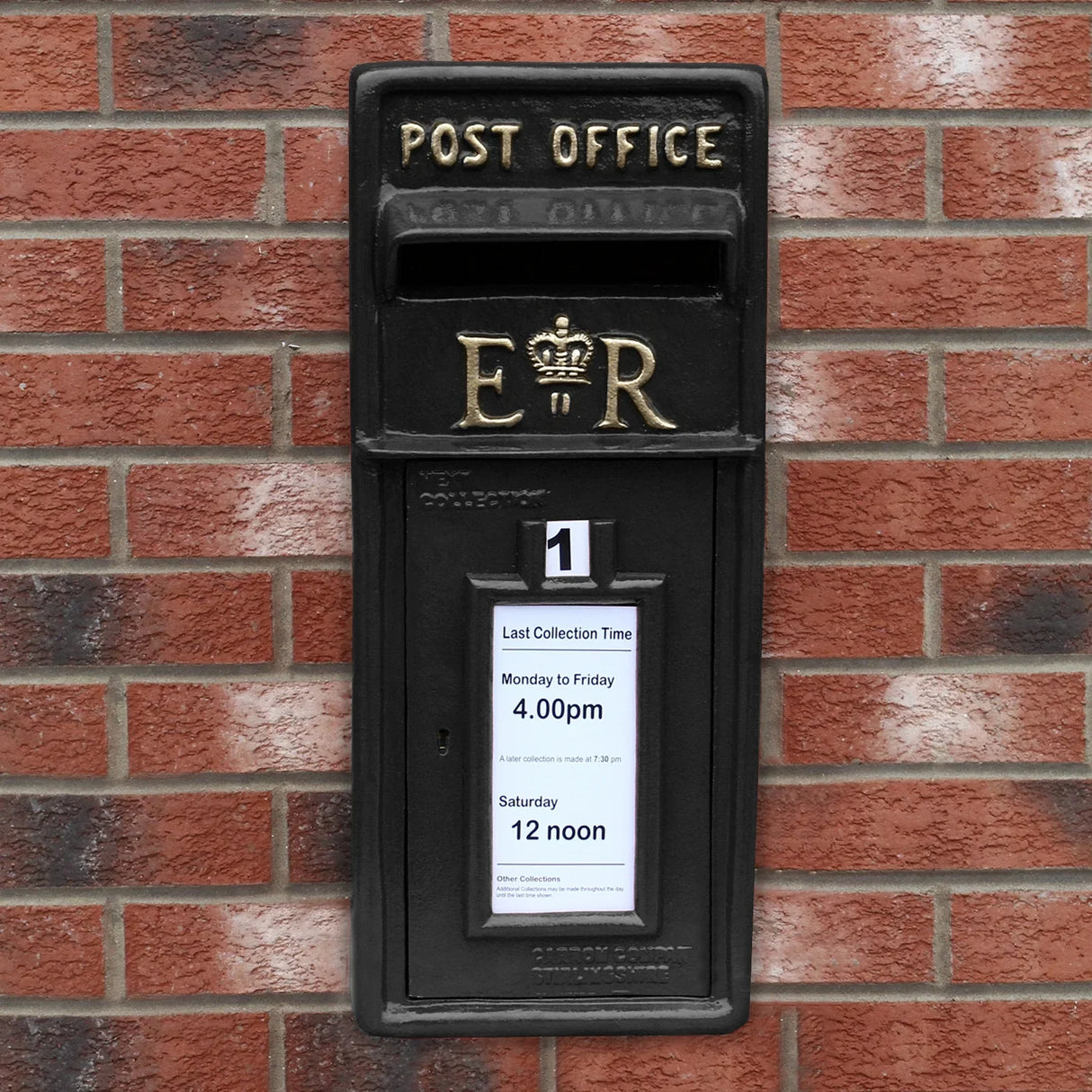 Black Royal Mail post box mounted on a brick wall, featuring gold lettering, collection times displayed, and a classic design