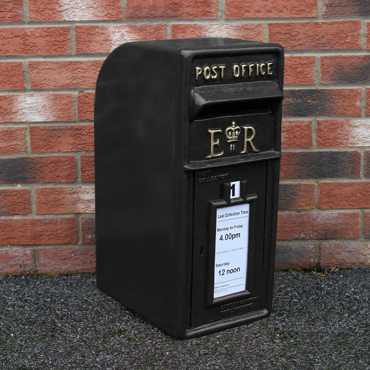 Black Royal Mail post box with gold lettering, featuring a crown emblem, set against a brick wall, showcasing collection times