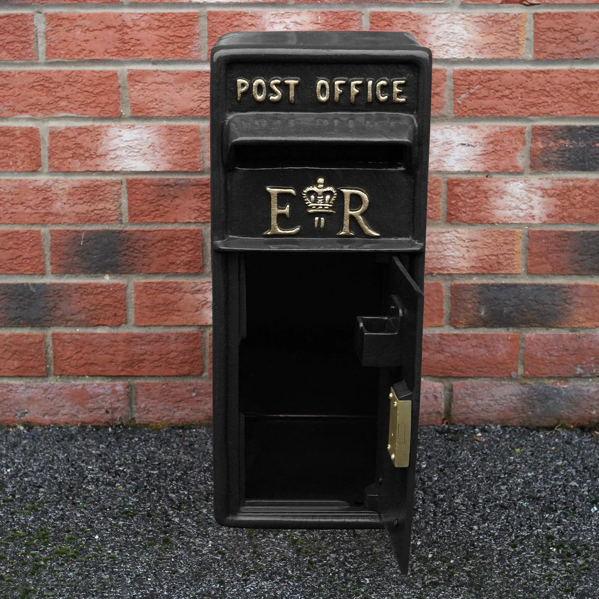 Black Royal Mail post box with gold lettering, featuring a classic design and an open door, set against a brick wall background