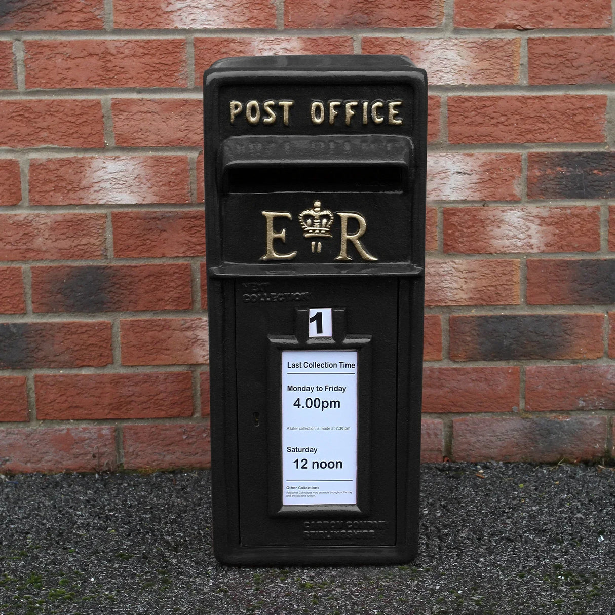 Black Royal Mail post box with gold lettering, featuring collection times on a white notice, set against a brick wall background