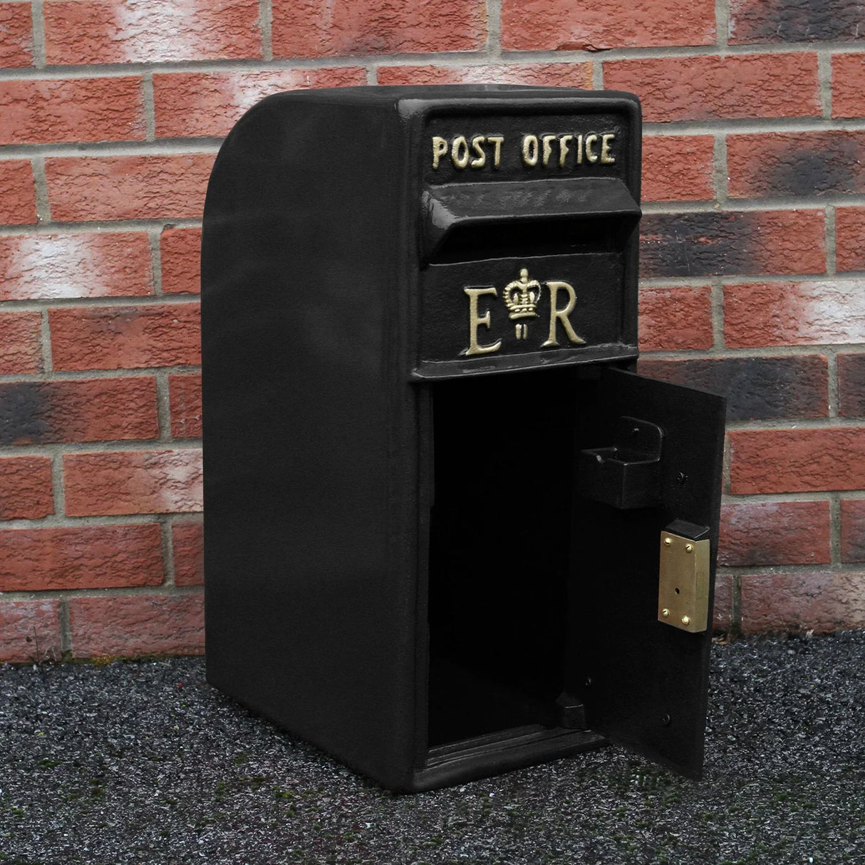 Black Royal Mail post box with gold lettering, featuring a curved top, open door, and classic design against a brick wall background