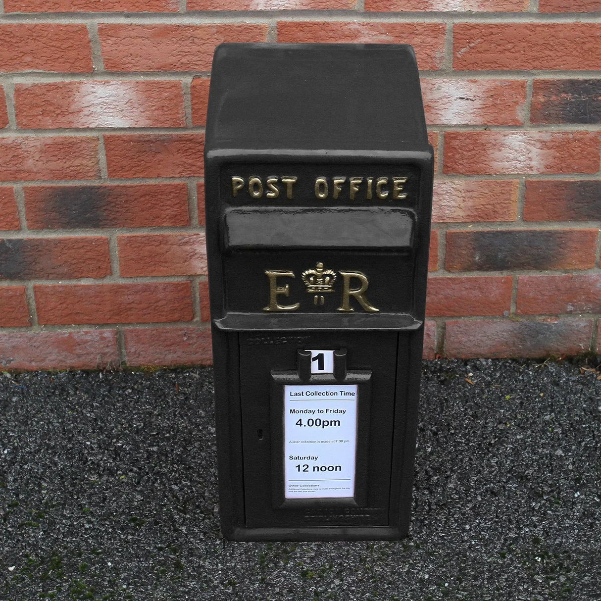 Black Royal Mail post box with gold lettering, featuring a collection time notice, set against a brick wall background