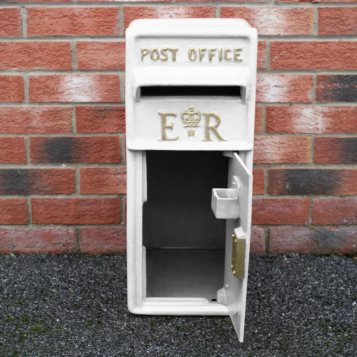 White Royal Mail post box with gold lettering, featuring a decorative crown and an open door, set against a brick wall background