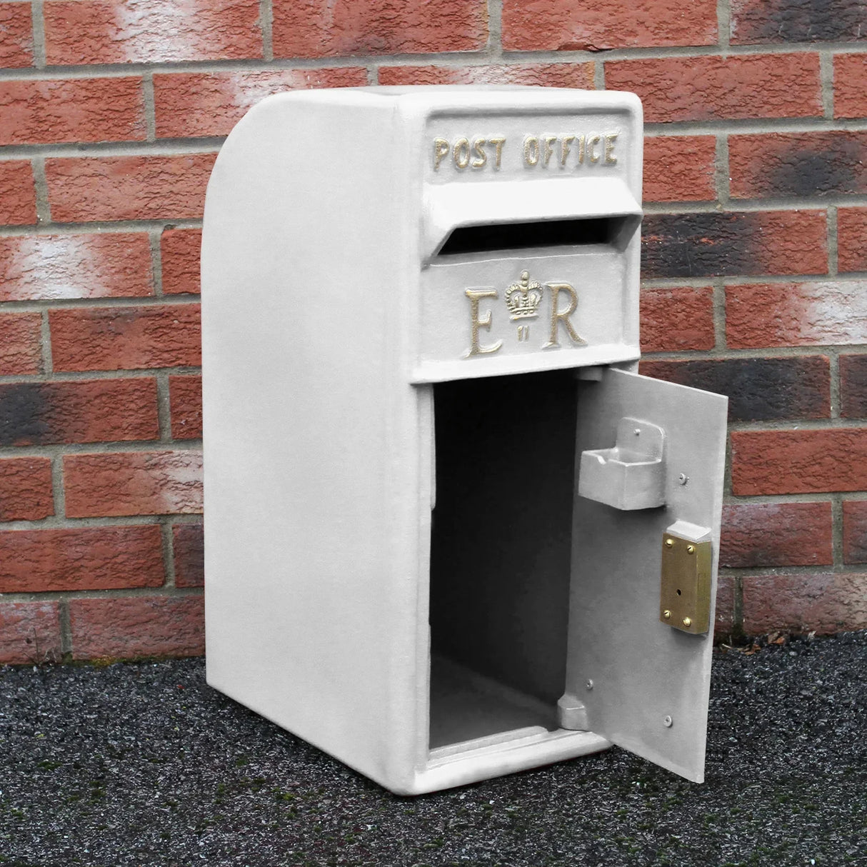 White Royal Mail post box with stand, featuring gold "ER" insignia, textured surface, and open door against a brick wall backdrop