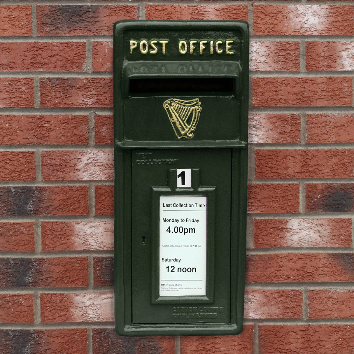 Green Irish post box mounted on a brick wall, featuring a gold harp emblem and a white collection time notice attached