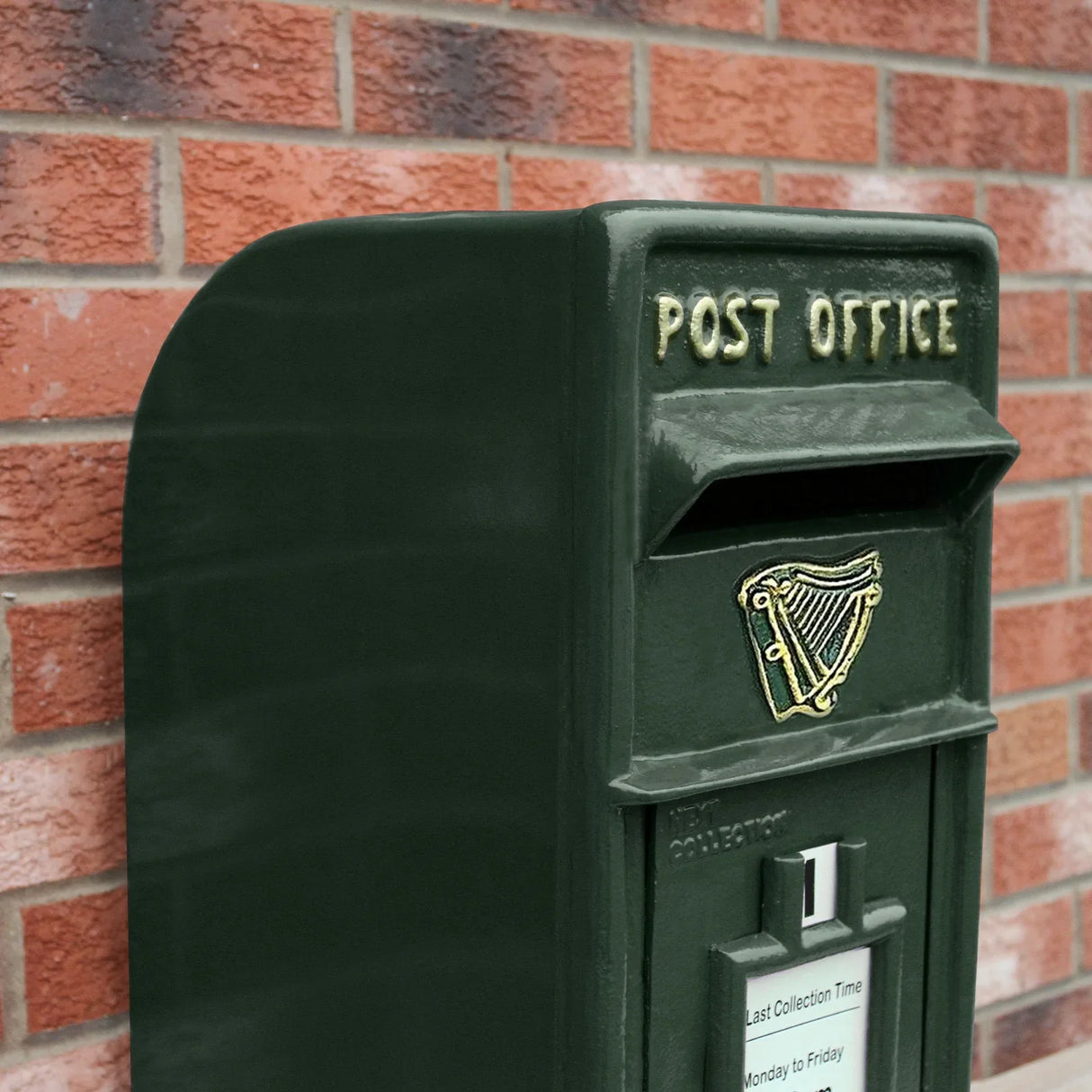 Green Irish post box with a curved top, gold harp emblem, and collection time details, set against a brick wall background