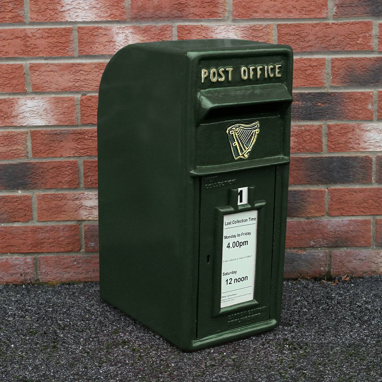 Green Irish post box with a curved top, textured surface, and gold harp emblem, set against a brick wall background
