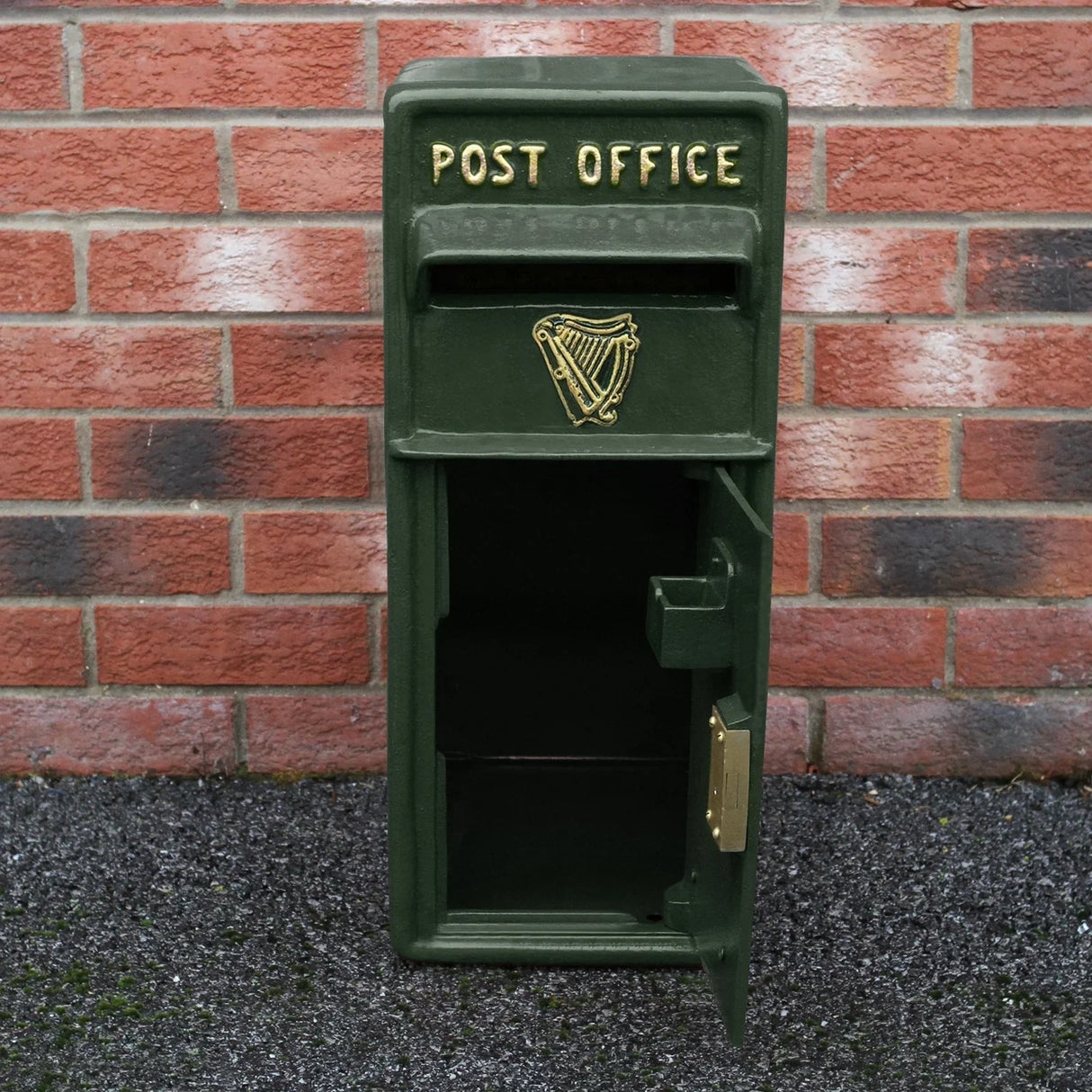 Vintage green Irish post box with gold lettering, featuring a harp emblem and an open front for mail collection against a brick wall
