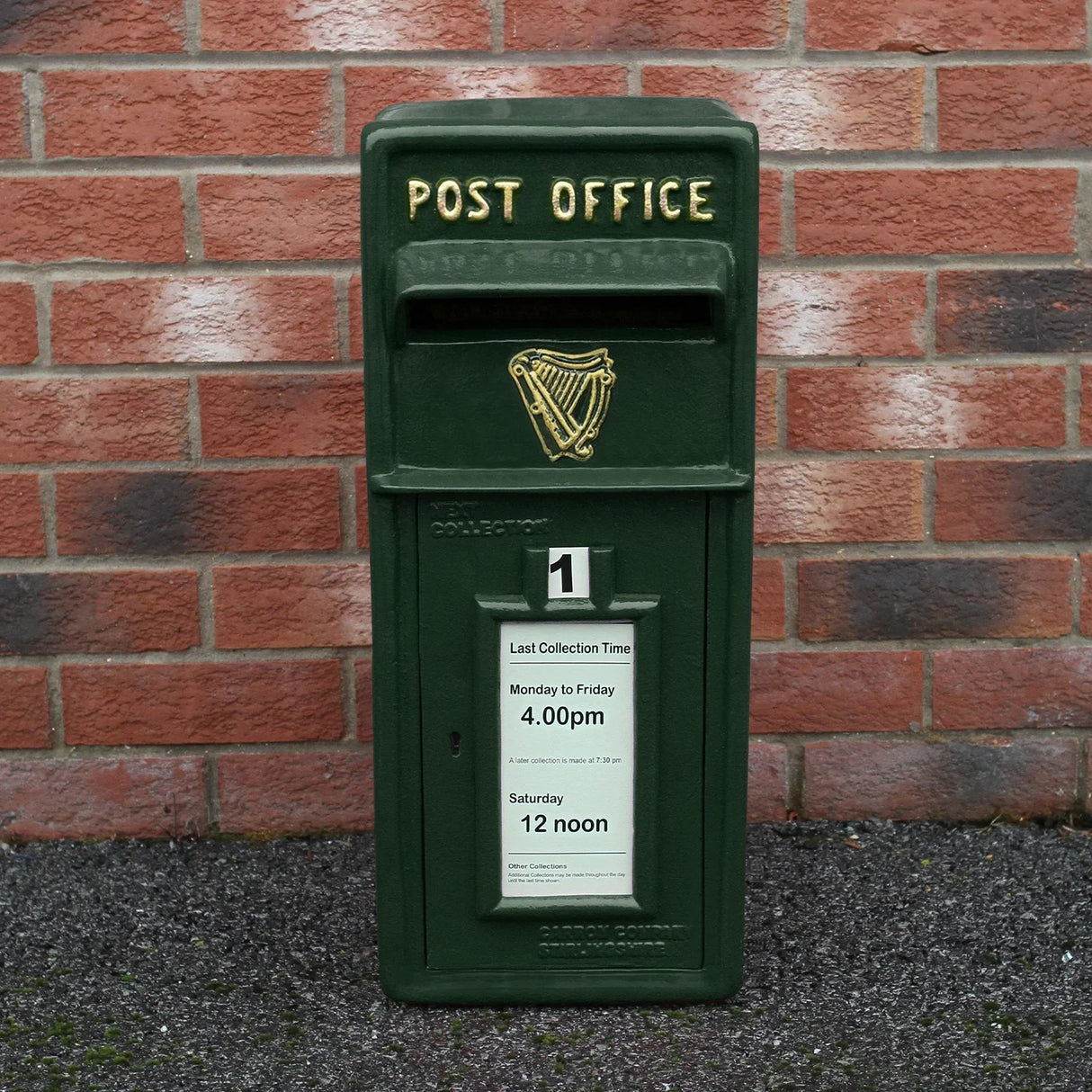 Green Irish post box with gold lettering, featuring a harp emblem and collection times displayed on a white notice against a brick wall