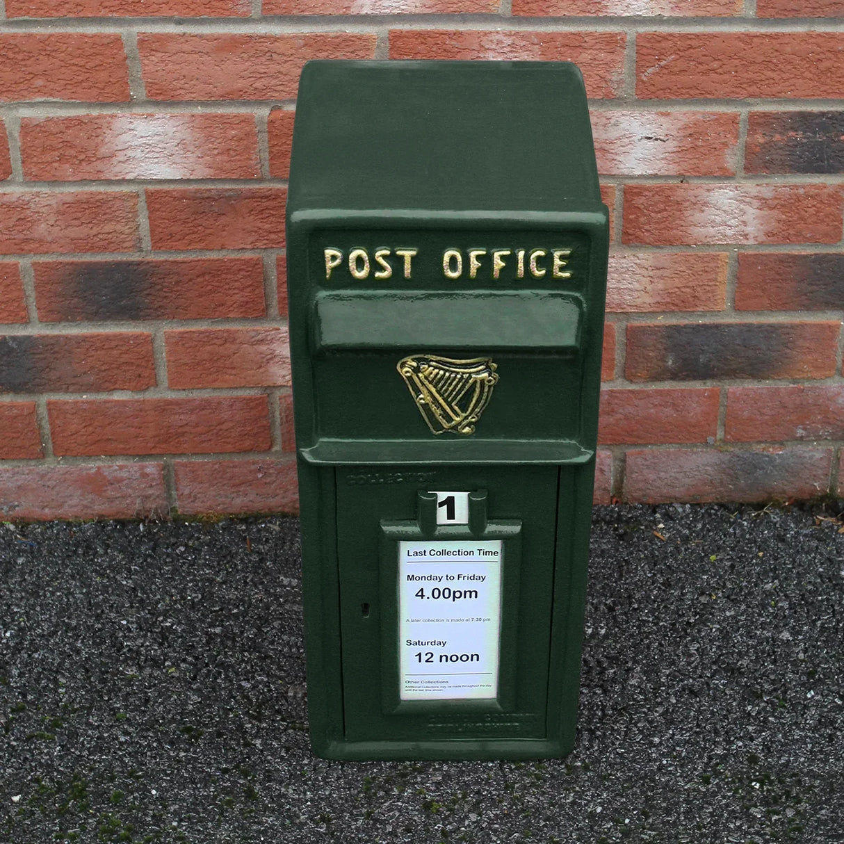 Green Irish post box with a textured finish, gold lettering, and collection times displayed on a white sign against a brick wall
