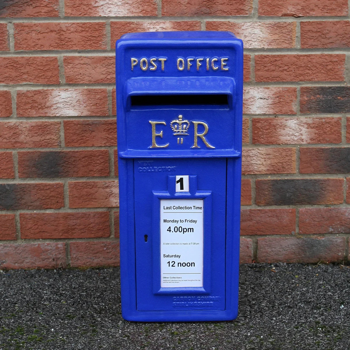 Bright blue Scottish post box with gold lettering, featuring a collection time notice, set against a brick wall background
