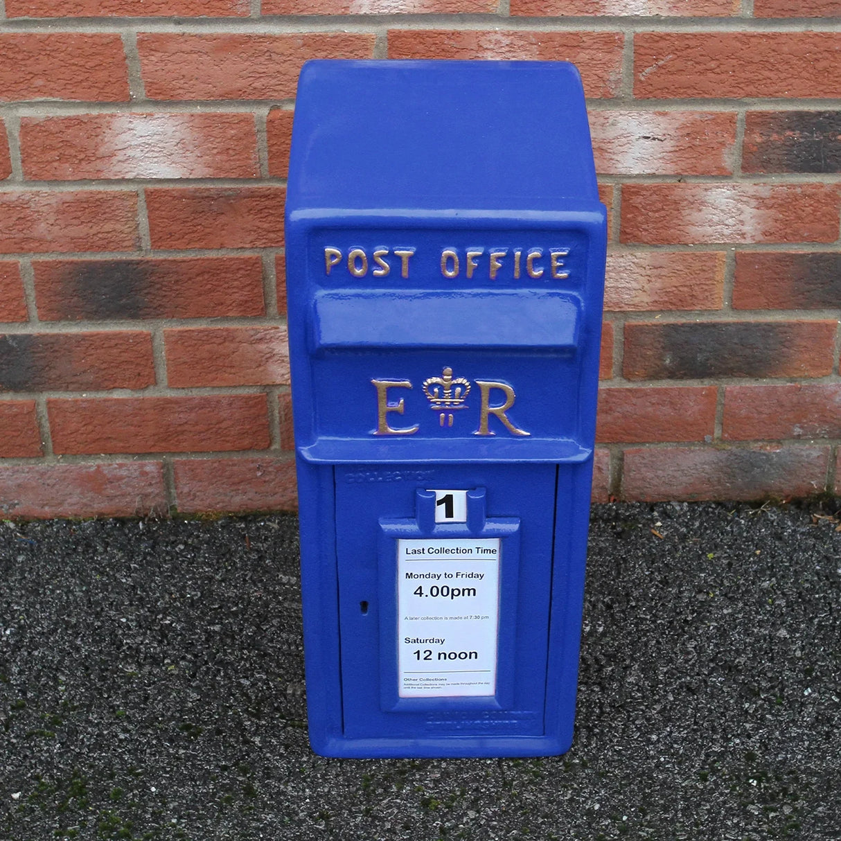 Bright blue Scottish post box with gold lettering, featuring a collection time notice and a crown emblem, set against a brick wall