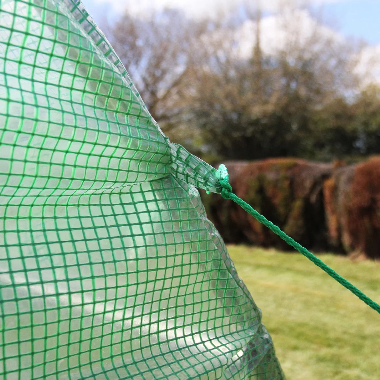 Close-up of the green mesh material of a Polytunnel 19mm 4m x 2m, showcasing its durable texture and secure tie points against a blurred garden backdrop