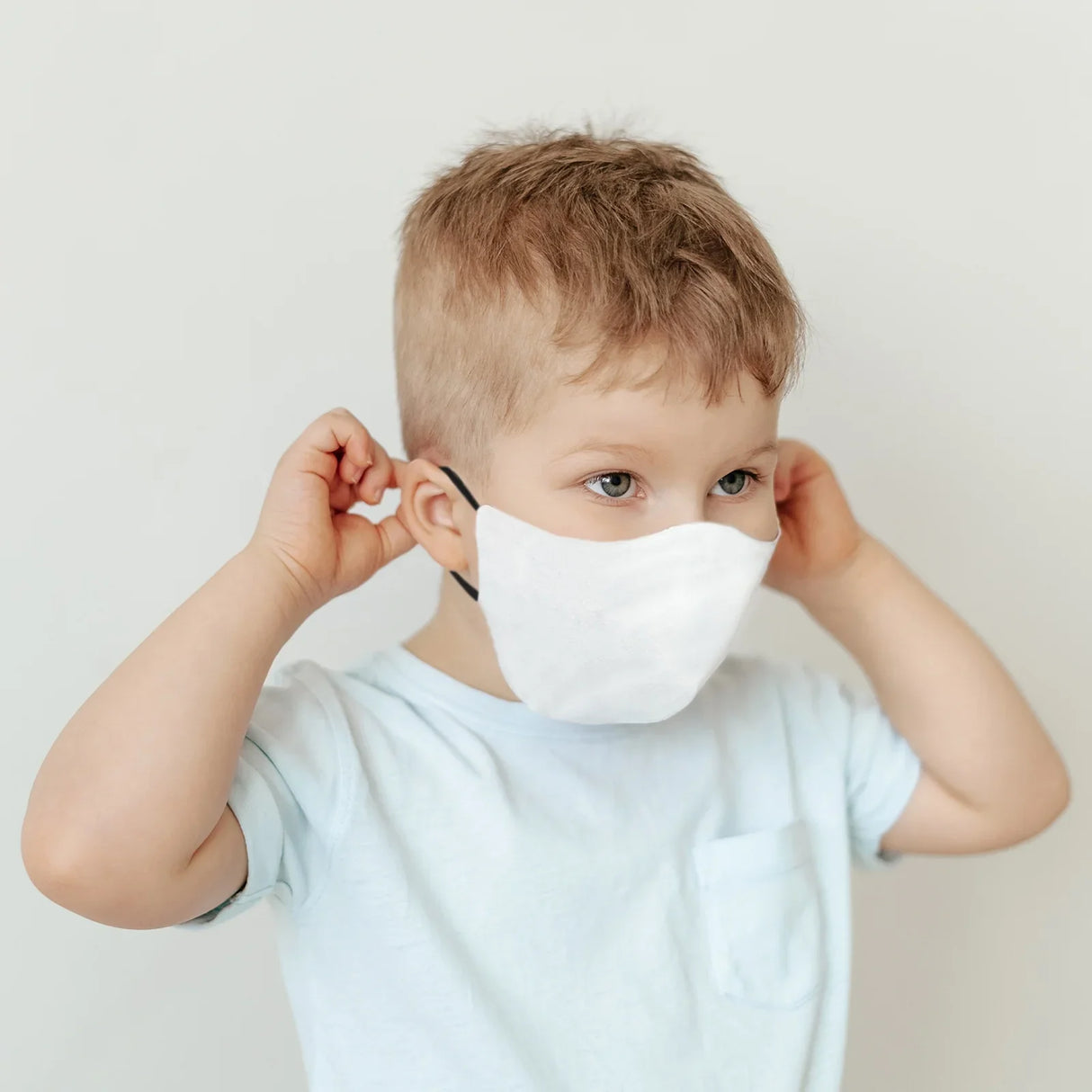 Child wearing a white face mask, adjusting it with both hands, showcasing a soft fabric texture and comfortable fit for kids