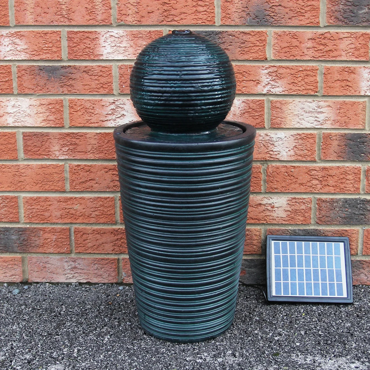 Round ball solar water feature with textured dark green finish, set atop a cylindrical base, beside a solar panel on a brick background