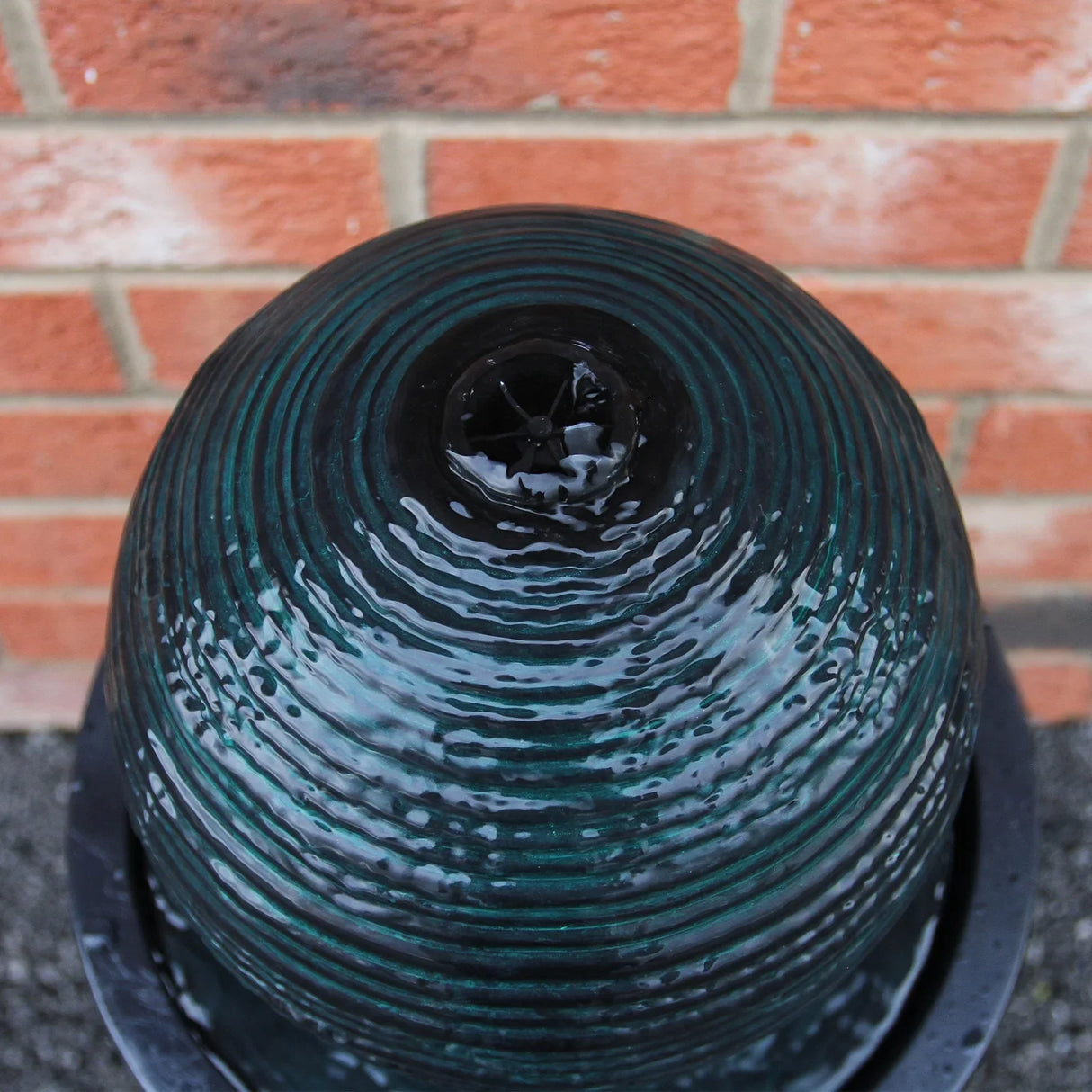 Round ball solar water feature with a textured dark green surface, gently flowing water, and a sleek black base against a brick wall