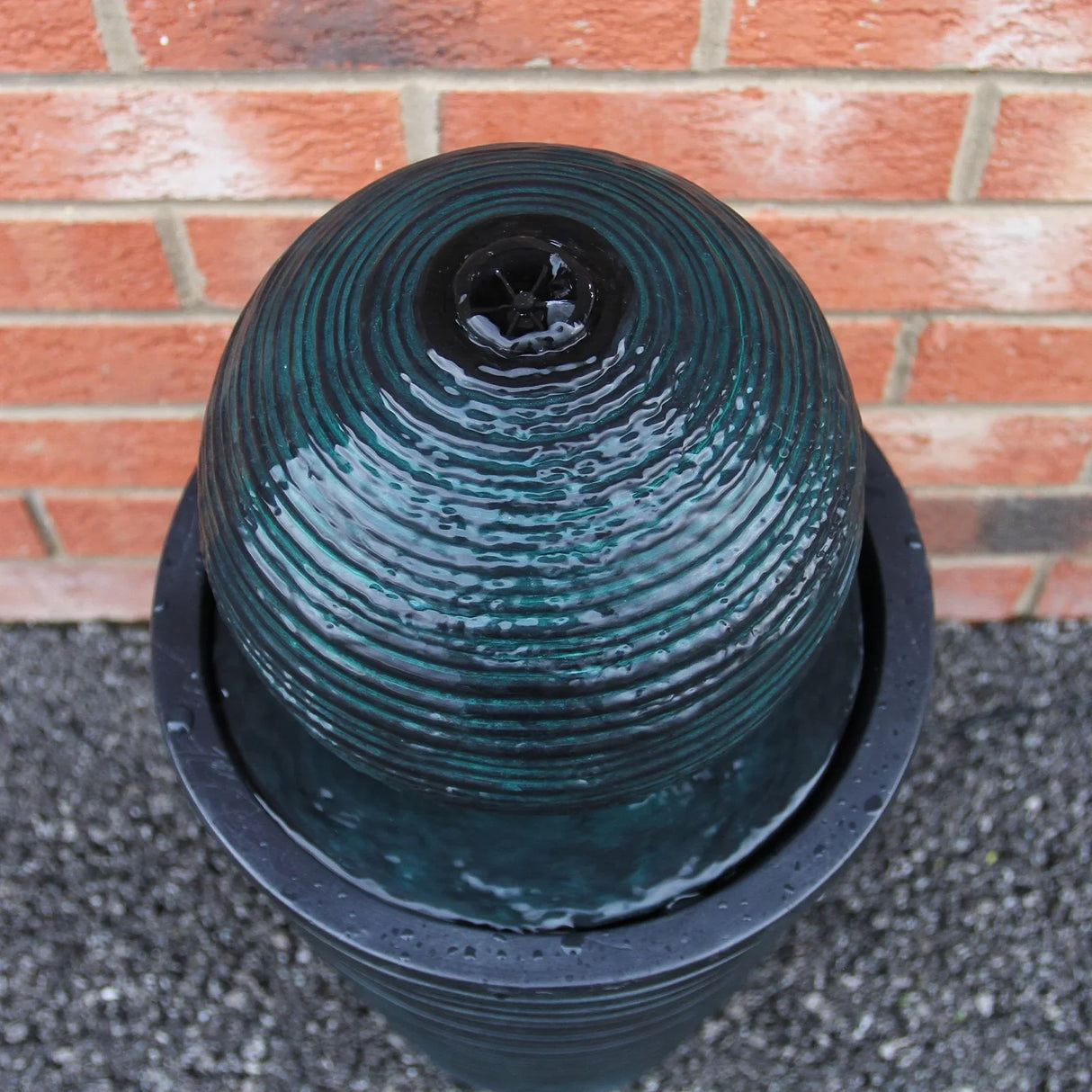 Round ball solar water feature with textured dark green finish, gently flowing water, and a sleek, modern design against a brick background