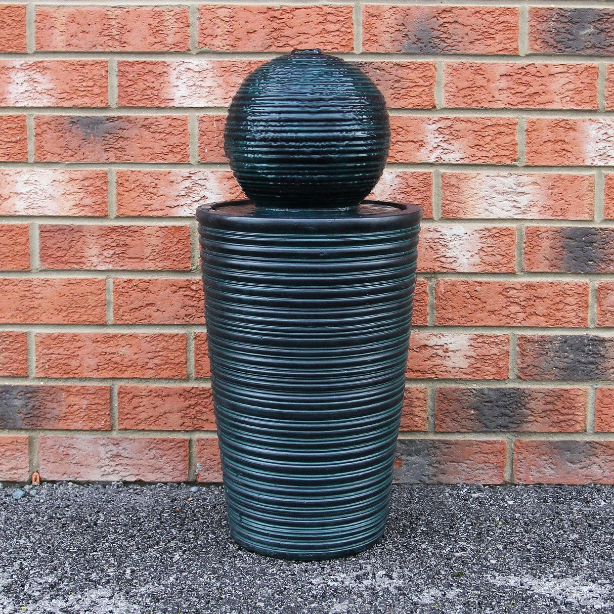 Round ball solar water feature with textured dark green finish, featuring a spherical top and cylindrical base against a brick wall