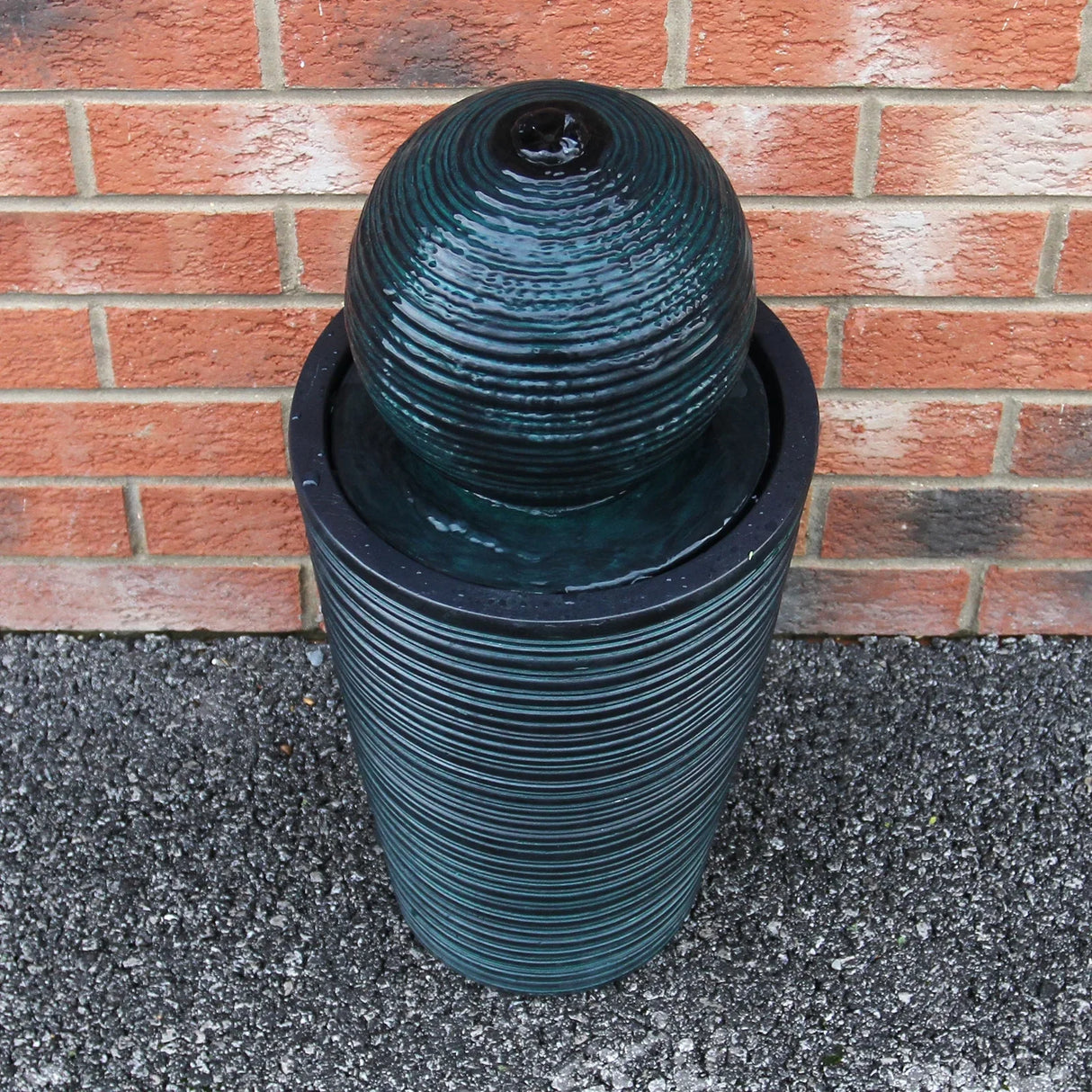 Round ball solar water feature with textured dark green finish, cascading water from the top into a circular basin, set against a brick wall