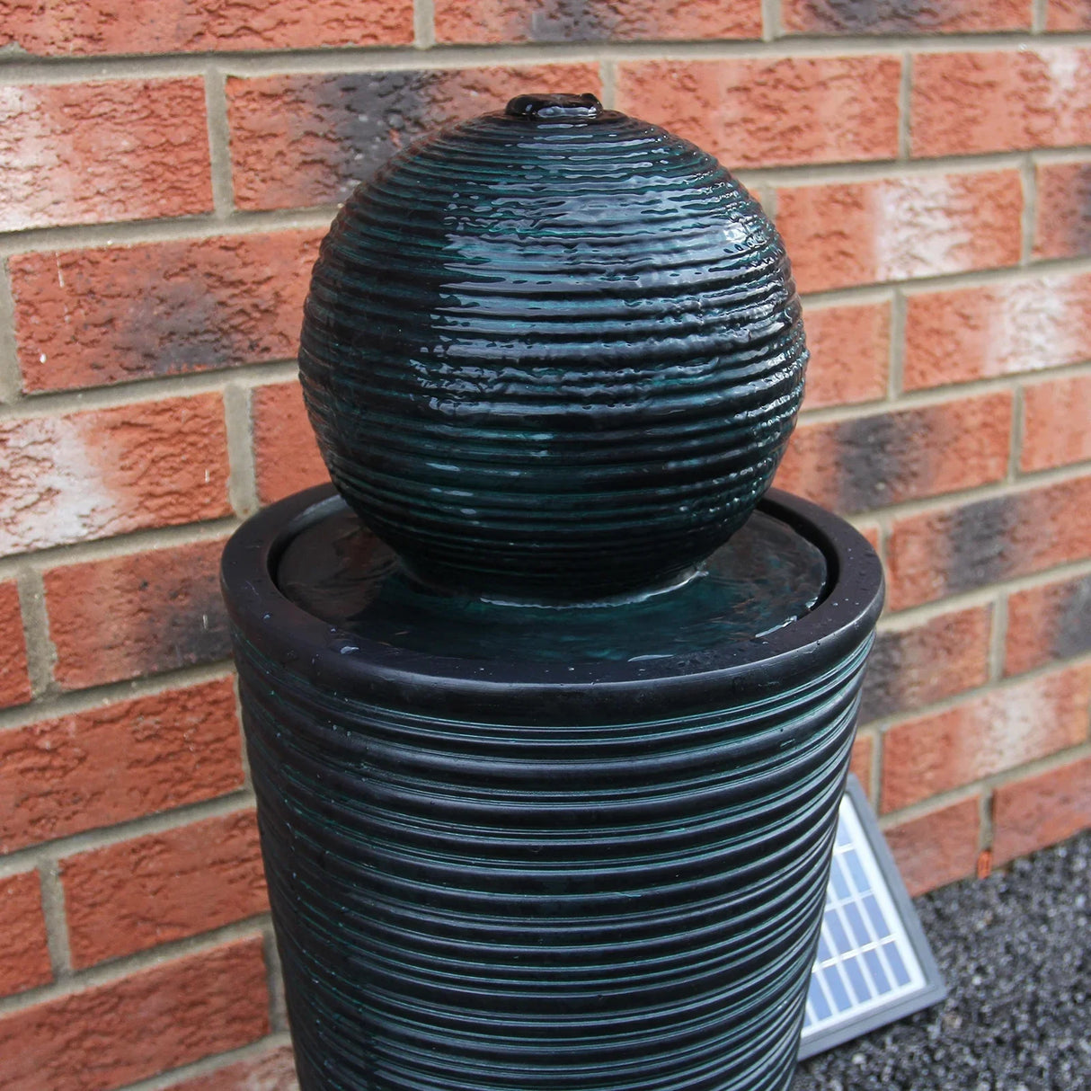 Round ball solar water feature with textured dark green finish, gently flowing water, and sleek cylindrical base against a brick wall