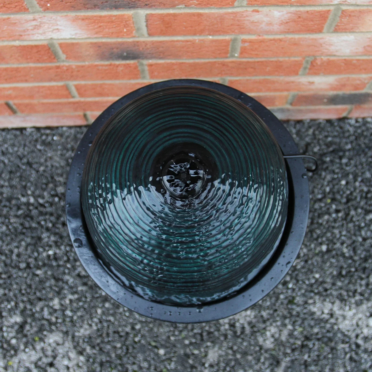 Round ball solar water feature with rippling dark green glass surface, set in a sleek black basin against a brick wall background