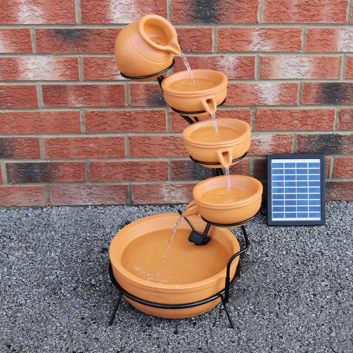 Terracotta 4 tier spilling bowls water feature with cascading water, black metal stand, and solar panel against a brick wall