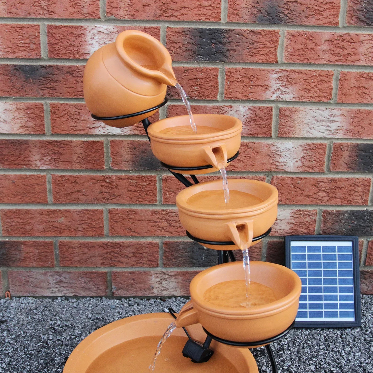 Terracotta 4 tier spilling bowls water feature with flowing water, rustic pots, and a solar panel against a brick wall
