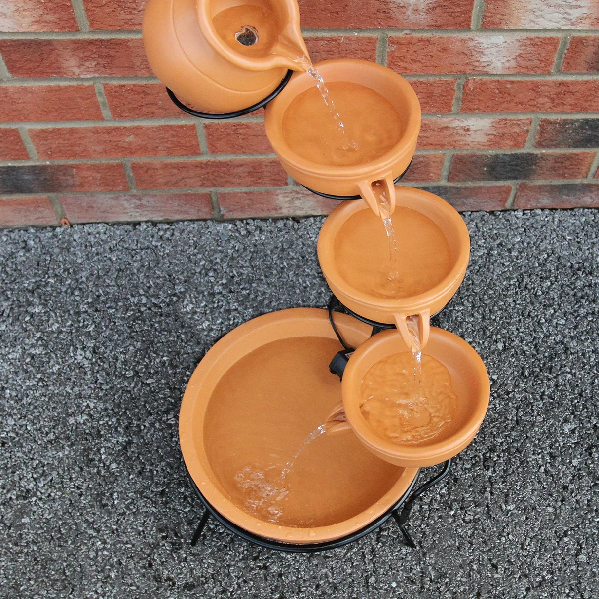 Terracotta 4 tier spilling bowls water feature with cascading water, earthy tones, and a sturdy black metal frame on a gravel surface