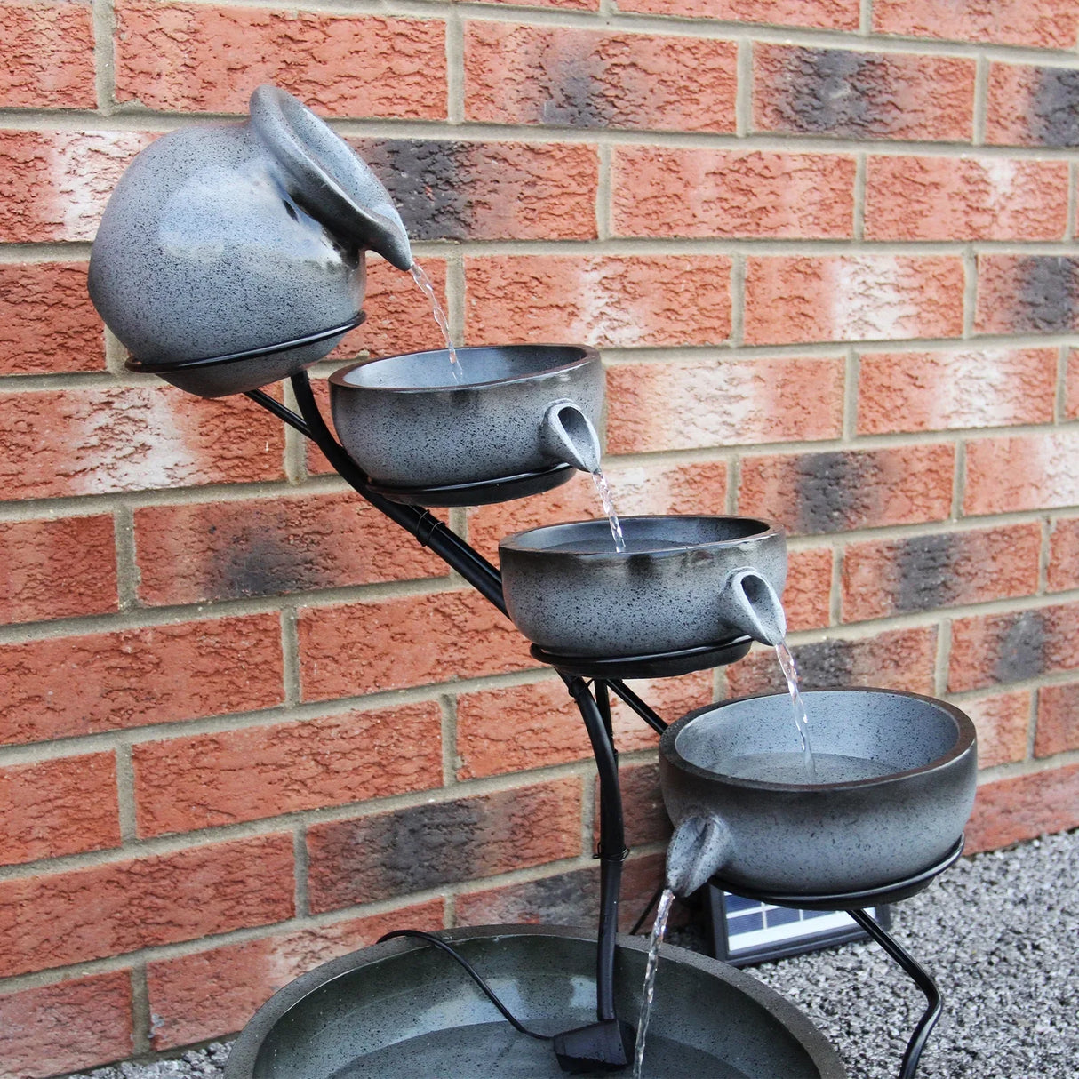 Grey 4 tier spilling bowls water feature with textured bowls, cascading water, and a sleek black frame against a brick wall