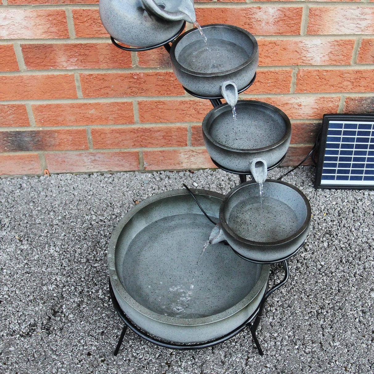 Grey 4 tier spilling bowls water feature with textured bowls, flowing water, and a solar panel, set against a brick wall