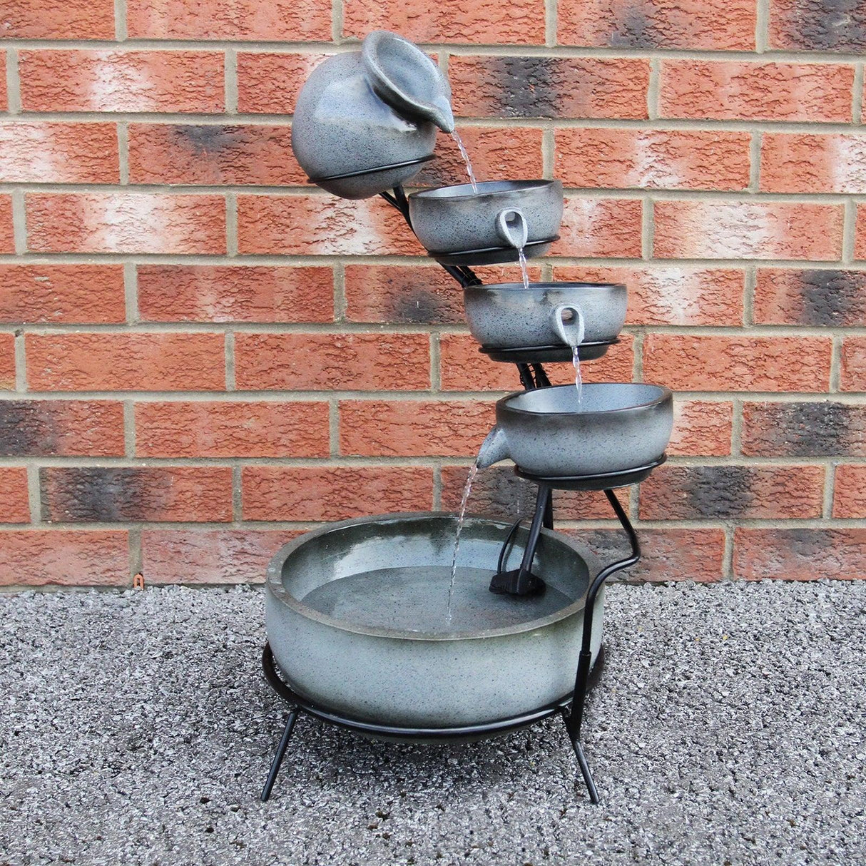 Grey 4 tier spilling bowls water feature with cascading water, textured ceramic bowls, and a sturdy black metal frame on a gravel surface