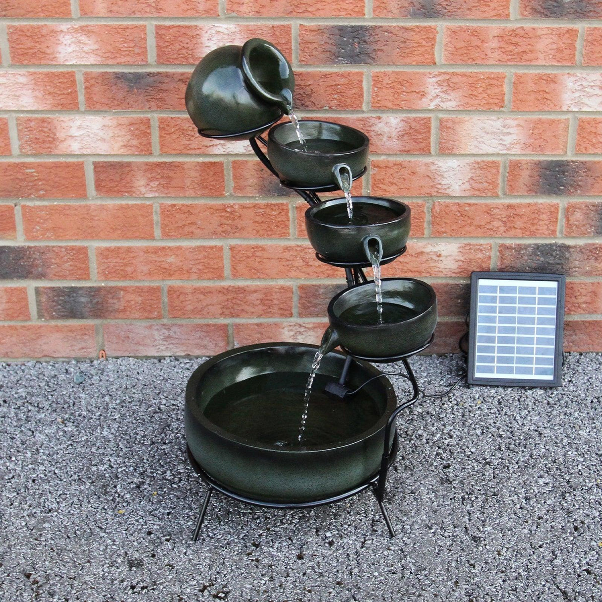 Green 4 tier spilling bowls water feature with a dark ceramic finish, cascading water, and a solar panel against a brick wall