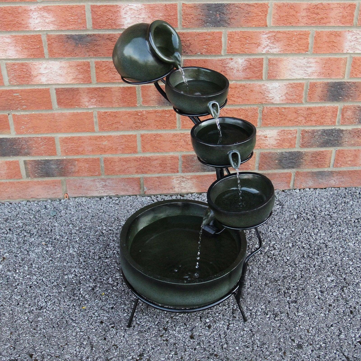 Green 4 tier spilling bowls water feature with smooth ceramic texture, cascading water, and a black metal stand against a brick wall