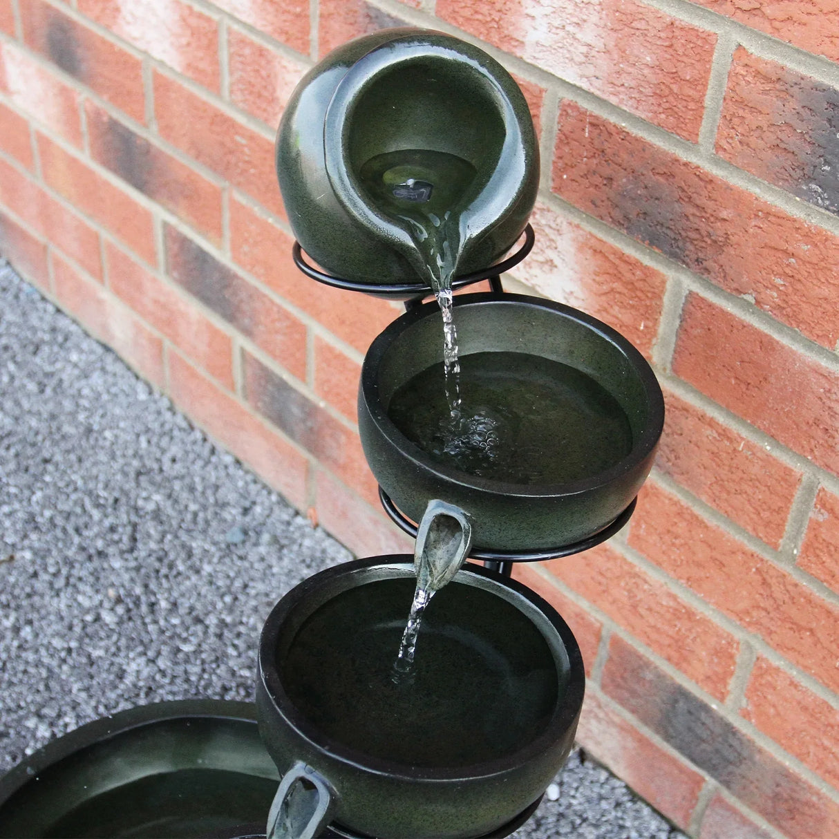 Green 4 tier spilling bowls water feature with smooth ceramic texture, cascading water, and a sleek black metal stand against a brick wall