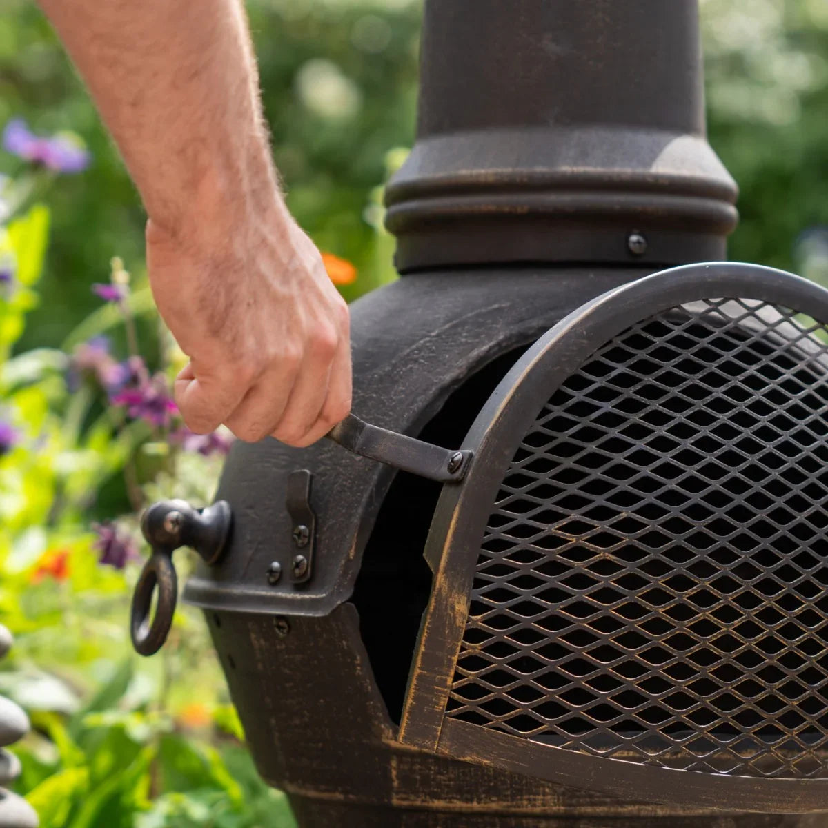 Rustic cast iron chiminea with a textured finish, featuring a mesh door and a hand opening it, surrounded by vibrant garden flowers