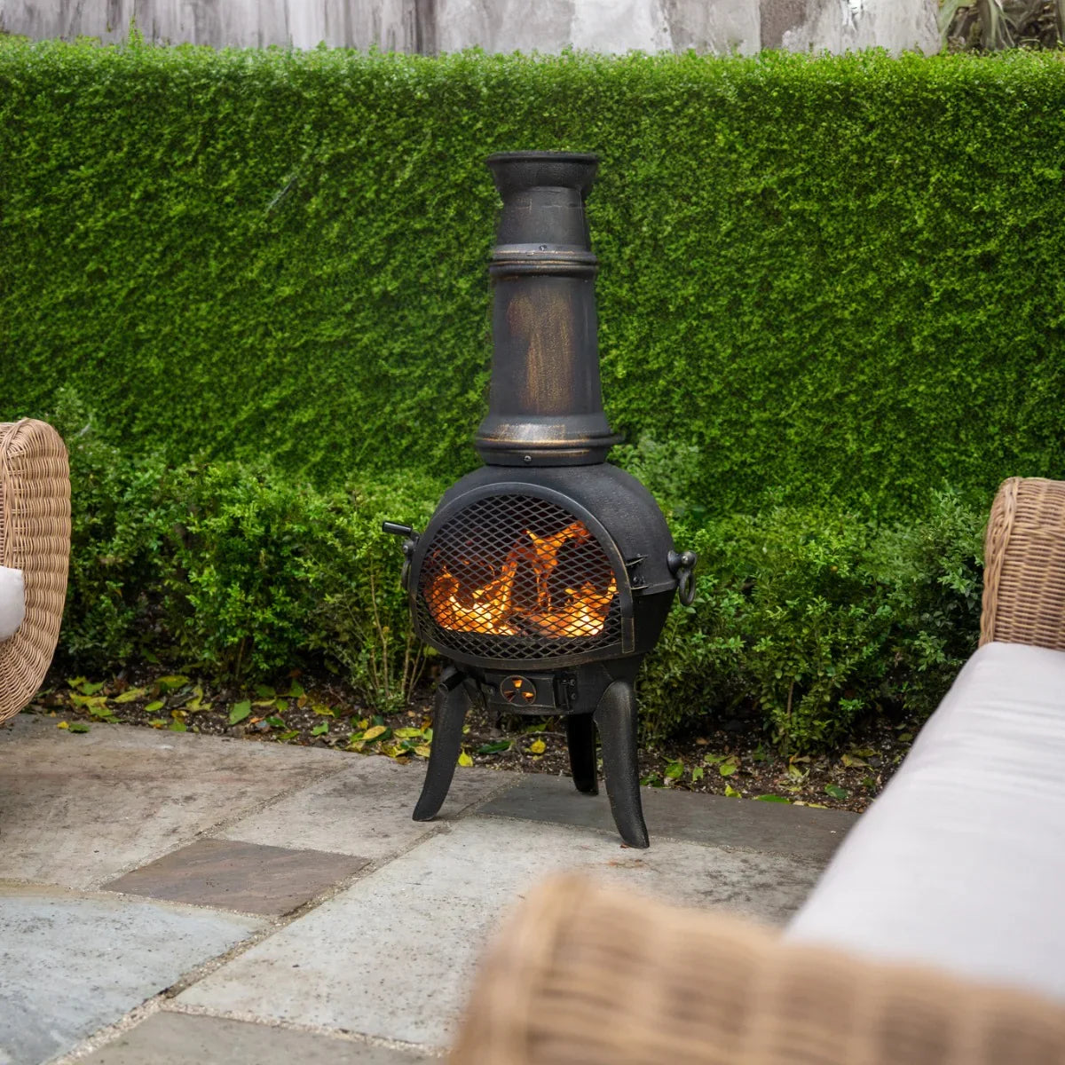 Black cast iron chiminea with flames, tall chimney, and mesh screen, surrounded by green hedges and outdoor seating