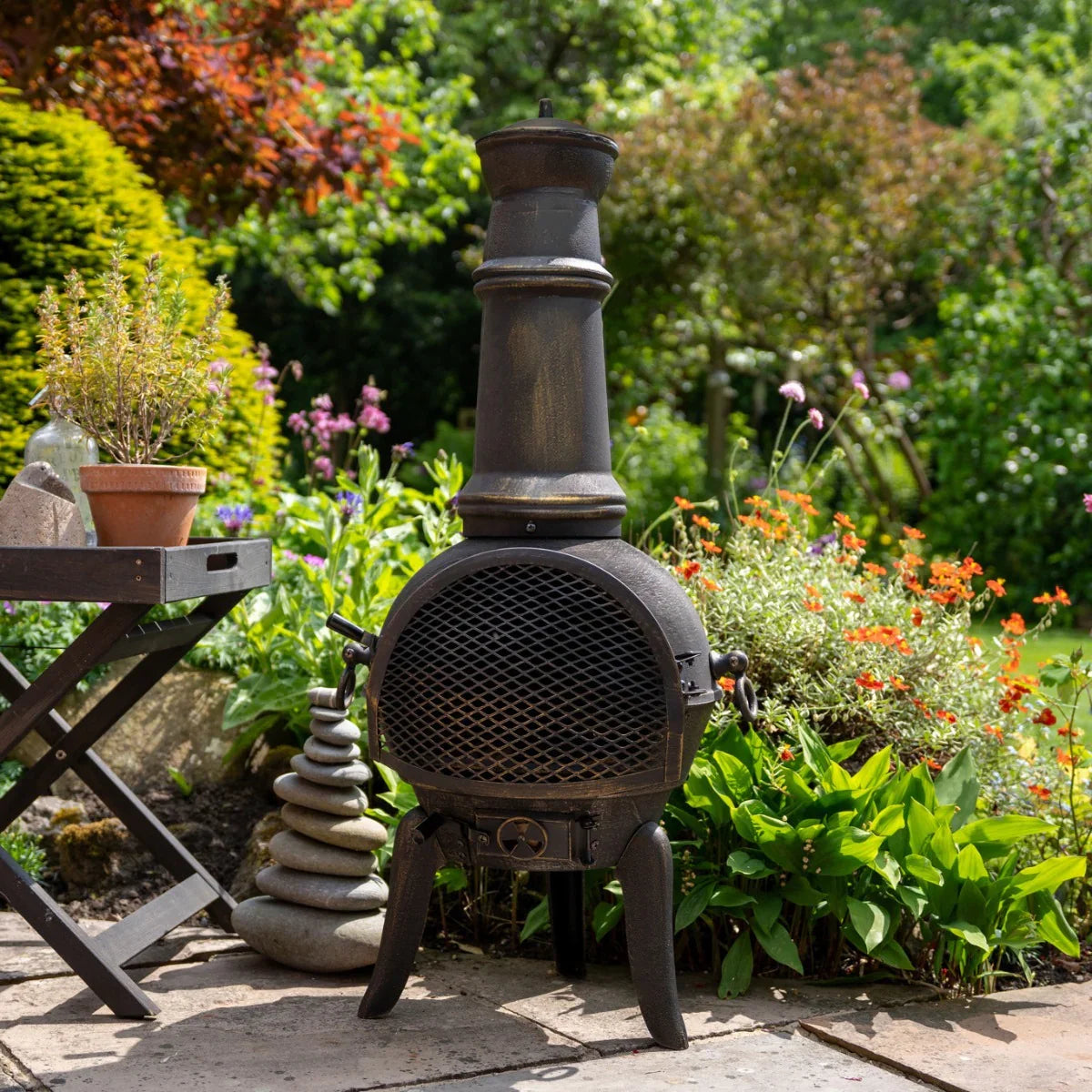Rustic cast iron chiminea with a tall chimney, black finish, mesh front, and surrounded by vibrant garden flowers and greenery