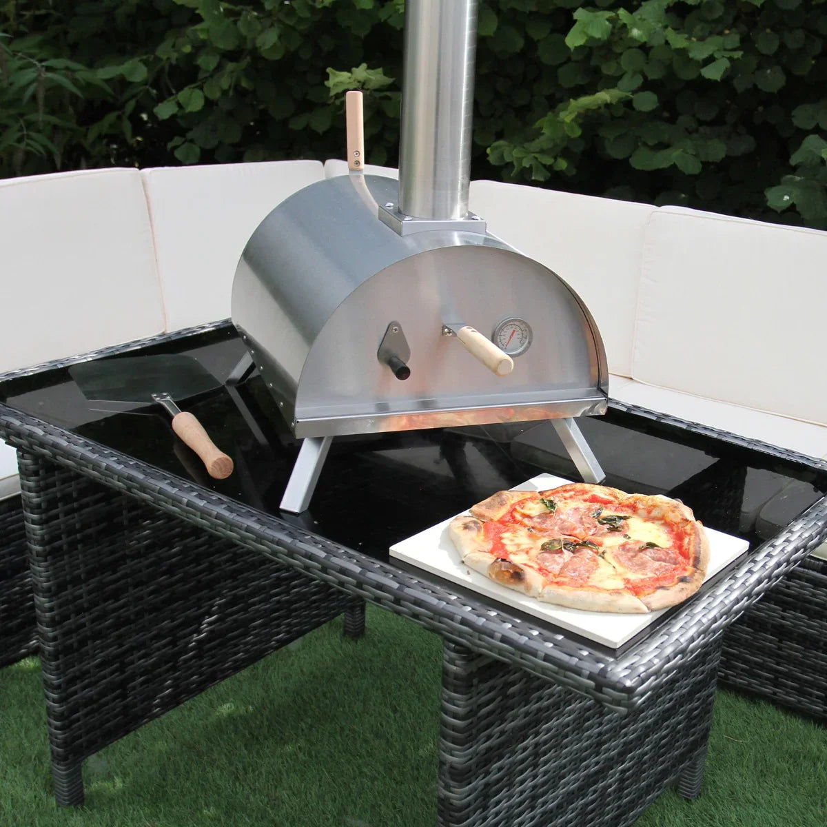 Outdoor table top pizza oven in stainless steel with a chimney, placed on a glass-top table beside a freshly baked pizza