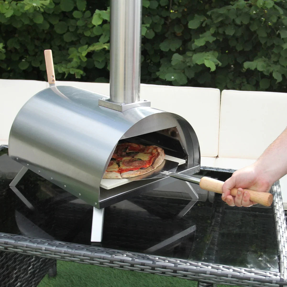 Stainless steel outdoor tabletop pizza oven with a chimney, featuring a pizza on a wooden peel and a sleek, modern design