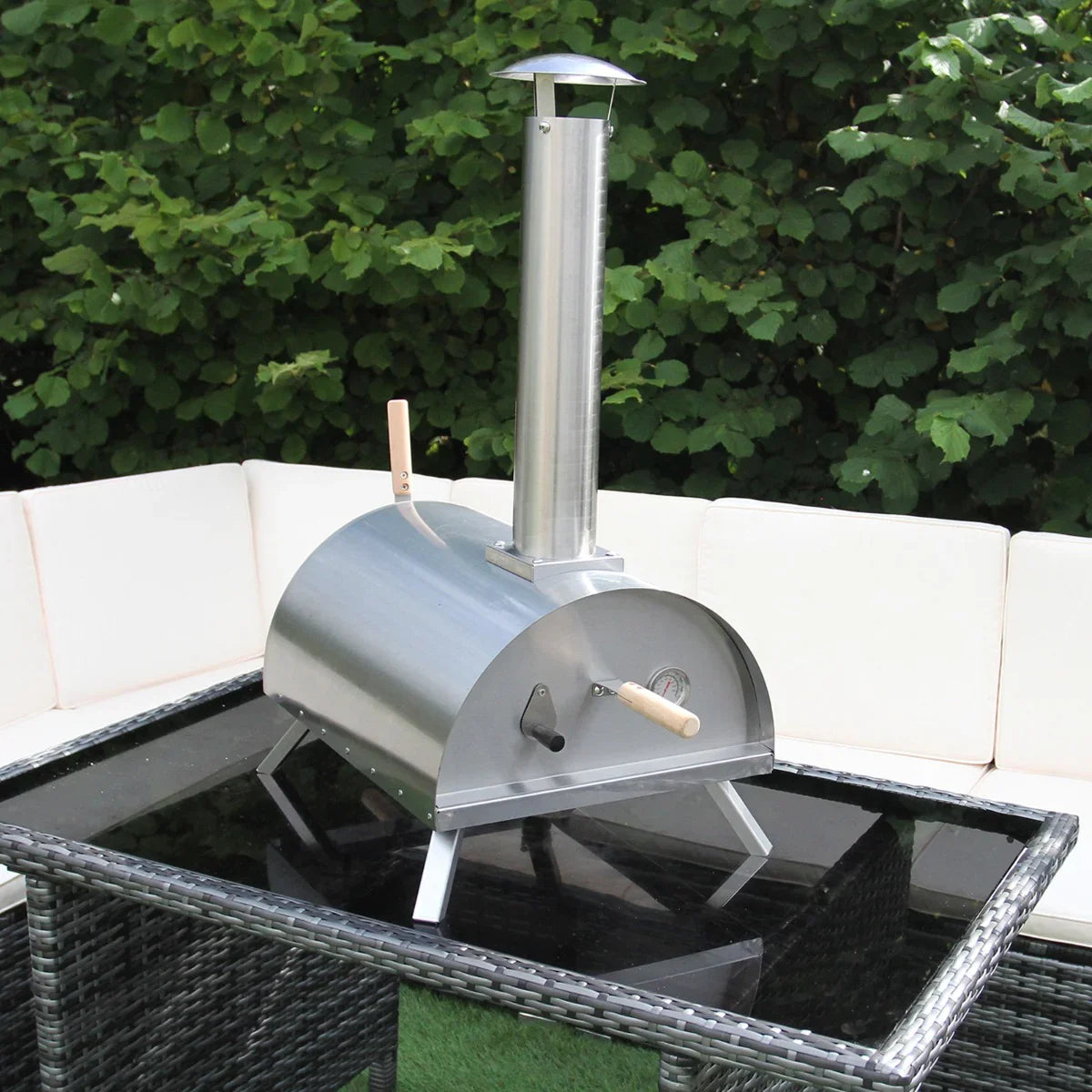 Outdoor table top pizza oven in stainless steel with a chimney, placed on a glass table surrounded by a cozy seating area