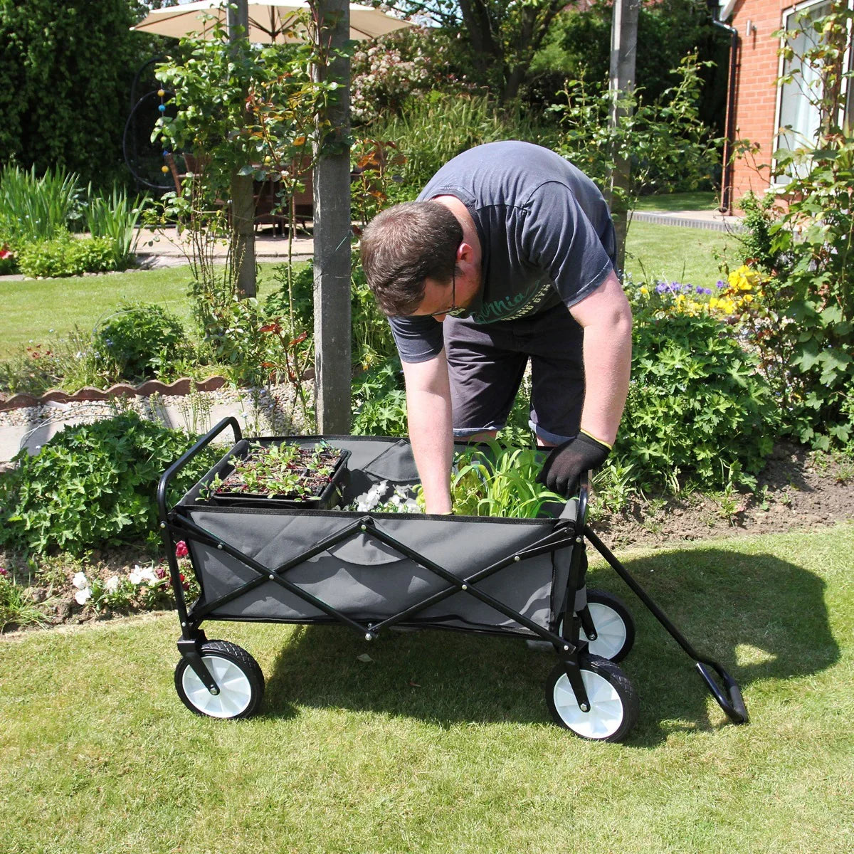 Foldable grey garden cart with black frame, filled with plants, on green lawn; user adjusting plants while gardening